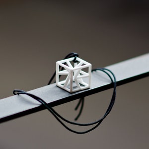 Op de afbeelding: Een witte geometrische ketting met een zwarte koord. De hanger is een kubus met een open ontwerp, waardoor een driedimensionaal effect ontstaat.
