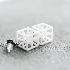 Op de afbeelding: Een paar witte geometrische kubus oorbellen met een delicaat draadontwerp. De oorbellen zijn gemaakt van een lichtgewicht materiaal en hebben een moderne, minimalistische esthetiek.