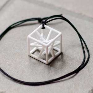 Op de afbeelding: Een witte geometrische ketting met een zwarte koord. De hanger is een kubus met een draadframe ontwerp.