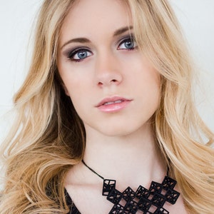 Może przedstawiać: Kobieta z długimi blond włosami nosi czarny geometryczny naszyjnik. Naszyjnik składa się z wielu małych, połączonych ze sobą sześcianów.