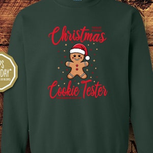 Könnte beinhalten: Grünes Christmas Cookie Tester Sweatshirt mit einem Lebkuchenmann, der einen Weihnachtsmann-Hut trägt. Der Text auf dem Sweatshirt lautet "Christmas Cookie Tester".