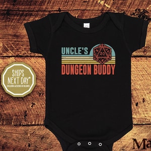 May include: Black baby bodysuit with the text "UNCLE'S DUNGEON BUDDY" in teal and red, with a D20 die graphic. The design is set against a background of yellow and teal stripes. The bodysuit has three snaps at the bottom.