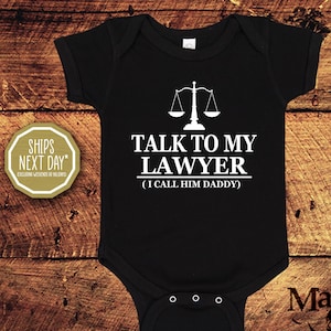 Puede incluir: Body de bebé negro con texto blanco que dice "TALK TO MY LAWYER (I CALL HIM DADDY)". Un gráfico blanco de una balanza de la justicia está encima del texto. El body es de manga corta y tiene tres corchetes en la parte inferior.