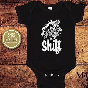 Könnte beinhalten: Schwarzer Baby-Body mit weißer Grafik eines Motocross-Fahrers und dem Wort "Shift". Das Design enthält den Text "Ohhhhhh" über dem Fahrer. Der Body hat drei Druckknöpfe am unteren Rand.