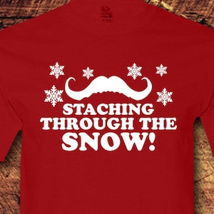 May include: Red t-shirt with a white mustache and snowflakes, and the text "Stacking through the snow!"
