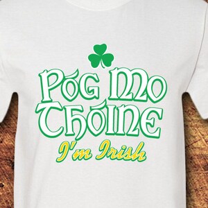 May include: White t-shirt with the text "Pog Mo Thoin" in green and yellow, topped with a green shamrock. The phrase "I'm Irish" is below the main text. The shirt has short sleeves and a crew neck.