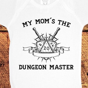 Puede incluir: Un body blanco para bebé con texto negro que dice "My Mom's the Dungeon Master" con una ilustración en blanco y negro de un dado de 20 caras y espadas cruzadas.