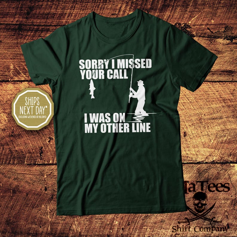 May include: Dark green t-shirt with white text: "Sorry I missed your call, I was on my other line." A graphic shows a fisherman and a fish. The shirt has a crew neck and short sleeves.