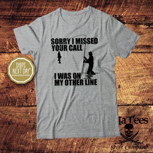 May include: A heather gray t-shirt with a fishing-themed graphic. The design features a fisherman, a fish, and the text "SORRY I MISSED YOUR CALL I WAS ON MY OTHER LINE" in black. The shirt has a crew neck and short sleeves.