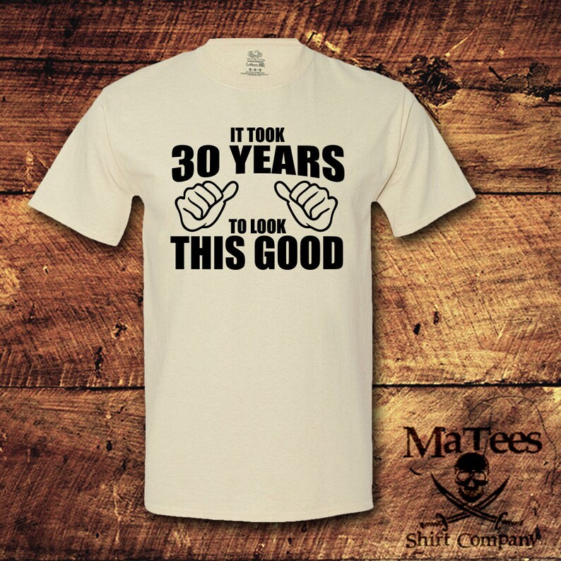 May include: A beige t-shirt with black text that reads "It took 30 years to look this good". The text is surrounded by two black hand drawn thumbs up.