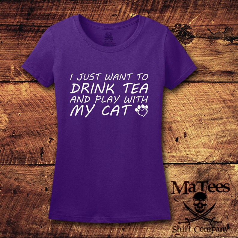May include: Purple short-sleeved t-shirt with white text that reads "I just want to drink tea and play with my cat" with a paw print and heart graphic.