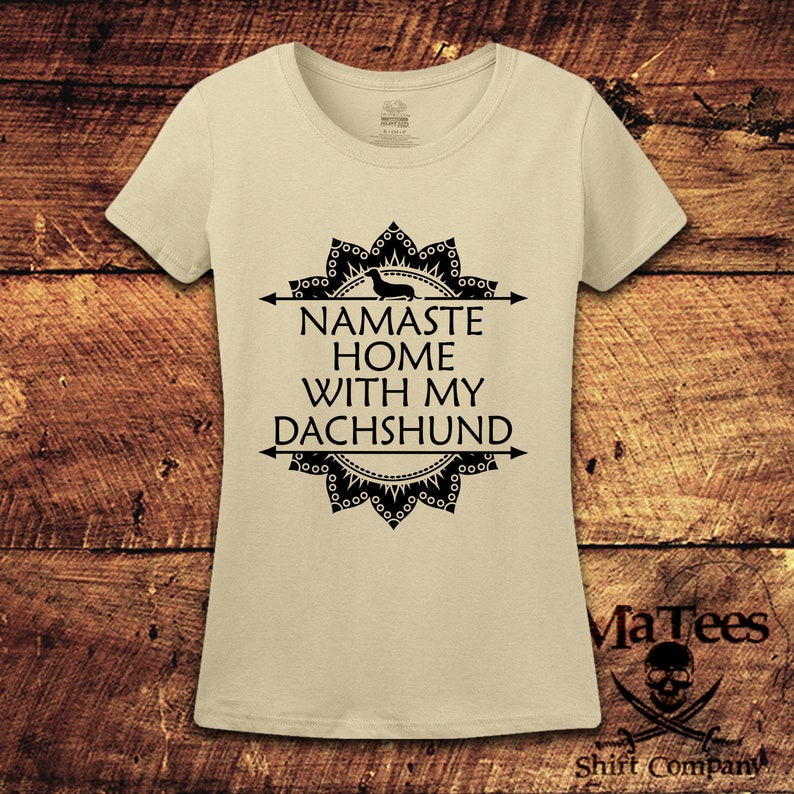 Op de afbeelding: Een beige t-shirt met een zwart mandala-ontwerp en de tekst "Namaste Home With My Dachshund".