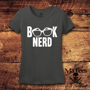 May include: Dark gray t-shirt with the words "BOOK NERD" in white, with glasses incorporated into the design. The shirt is displayed on a wooden background. The brand name "MaTees Shirt Company" is printed on the bottom right.