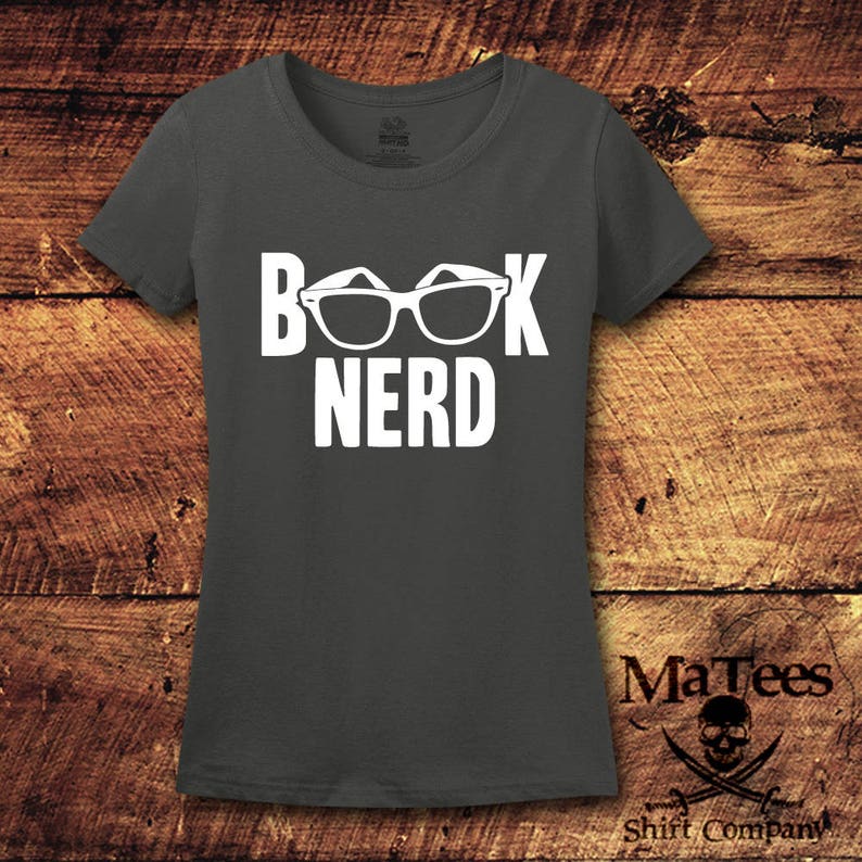 May include: Dark gray t-shirt with the words "BOOK NERD" in white, with glasses incorporated into the design. The shirt is displayed on a wooden background. The brand name "MaTees Shirt Company" is printed on the bottom right.
