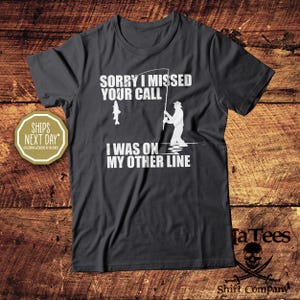 May include: Dark gray t-shirt with white text and graphics. The text reads "SORRY I MISSED YOUR CALL, I WAS ON MY OTHER LINE." A graphic shows a fisherman and a fish. The shirt is from TaTees Shirt Company.