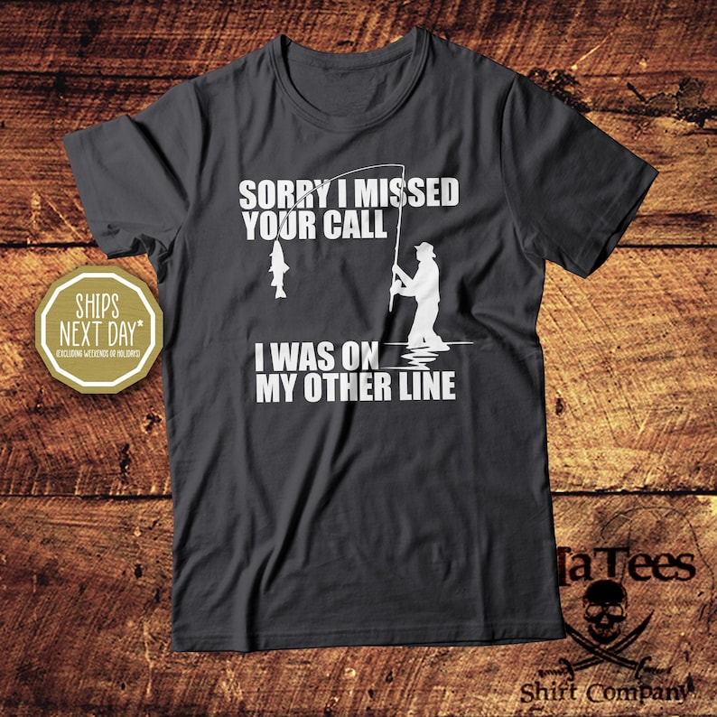May include: Dark gray t-shirt with white text and graphics. The text reads "SORRY I MISSED YOUR CALL, I WAS ON MY OTHER LINE." A graphic shows a fisherman and a fish. The shirt is from TaTees Shirt Company.