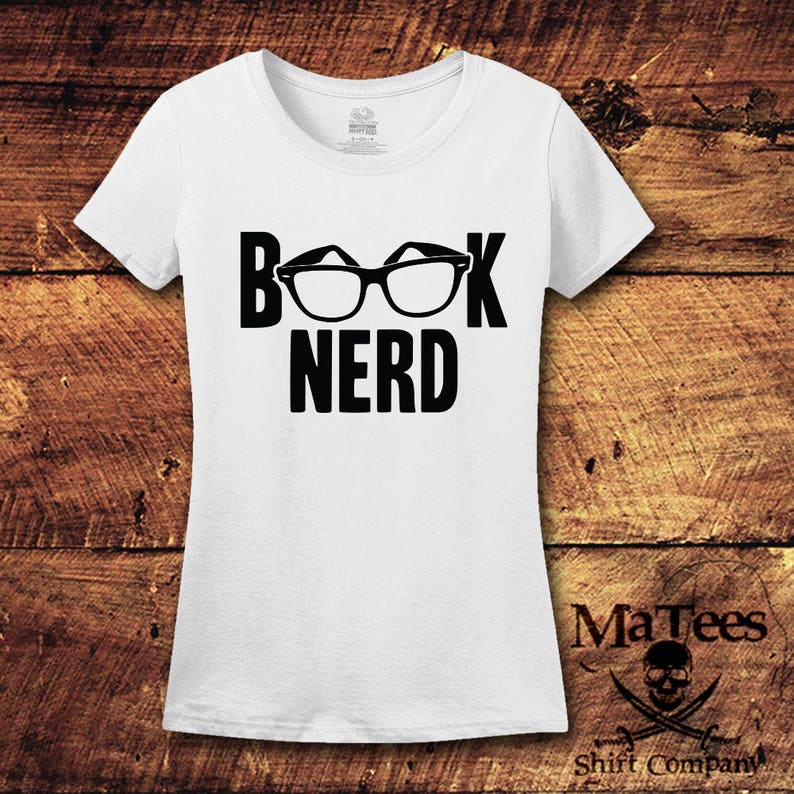 May include: White t-shirt with the words "BOOK NERD" in bold black letters. The word "BOOK" has a pair of black glasses in place of the "OO". The shirt is on a wooden background. The shirt is from MaTees Shirt Company.