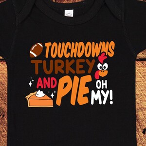 May include: Black baby onesie with the text "Touchdowns Turkey and Pie Oh My!" in orange, brown, and white. A football, a turkey, and a slice of pie are included in the design.