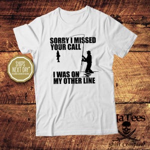 May include: White t-shirt with a black graphic of a fisherman and the text "SORRY I MISSED YOUR CALL I WAS ON MY OTHER LINE." The shirt also features a fish on the fishing line. The shirt is from the "Shirt Company".
