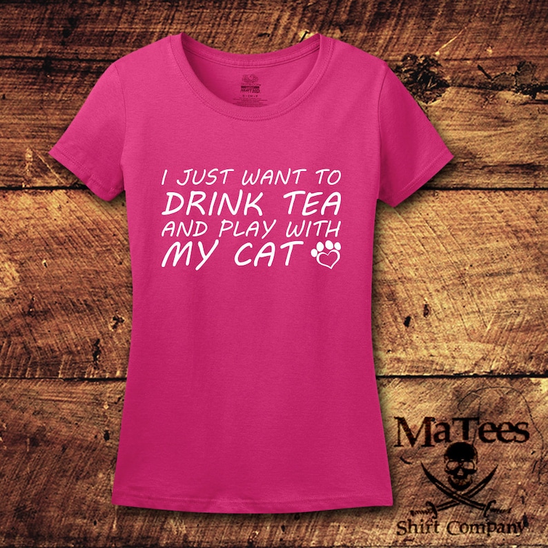 May include: Pink women's t-shirt with white text that reads "I just want to drink tea and play with my cat" with a paw print and heart graphic.