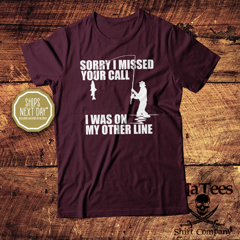 May include: A maroon t-shirt with white text and a graphic of a fisherman. The text reads "SORRY I MISSED YOUR CALL I WAS ON MY OTHER LINE". A fish is also depicted. The shirt is from Ta Tees Shirt Company.