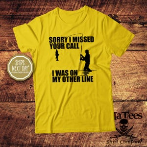 May include: Yellow t-shirt with black text and a fishing-themed graphic. The text reads "Sorry I missed your call, I was on my other line." A silhouette of a fisherman and a fish are also featured.