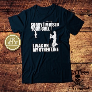 May include: Navy blue t-shirt with white text and a fishing-themed graphic. The text reads "SORRY I MISSED YOUR CALL I WAS ON MY OTHER LINE". A fisherman silhouette is shown with a fish and fishing rod. The shirt is from TaTees Shirt Company.