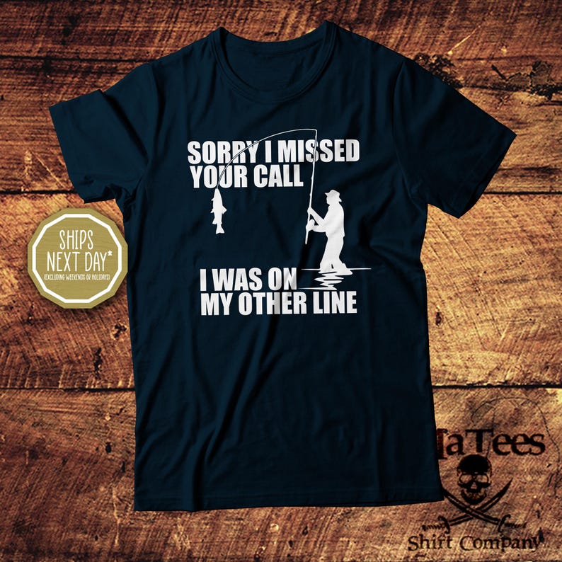 May include: Navy blue t-shirt with white text and a fishing-themed graphic. The text reads "SORRY I MISSED YOUR CALL I WAS ON MY OTHER LINE". A fisherman silhouette is shown with a fish and fishing rod. The shirt is from TaTees Shirt Company.
