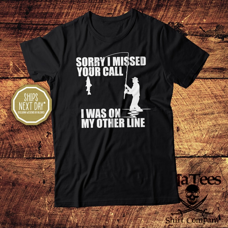 May include: Black t-shirt with the text "SORRY I MISSED YOUR CALL I WAS ON MY OTHER LINE" in white. A graphic of a fisherman and a fish are also in white. The shirt is from TaTees Shirt Company.