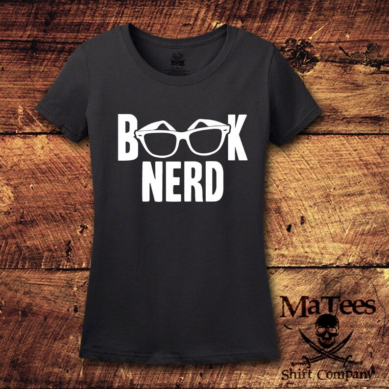 May include: Black t-shirt with the white text "BOOK NERD" and a graphic of eyeglasses. The shirt has a crew neck and short sleeves. The brand name "MaTees Shirt Company" is printed at the bottom right.