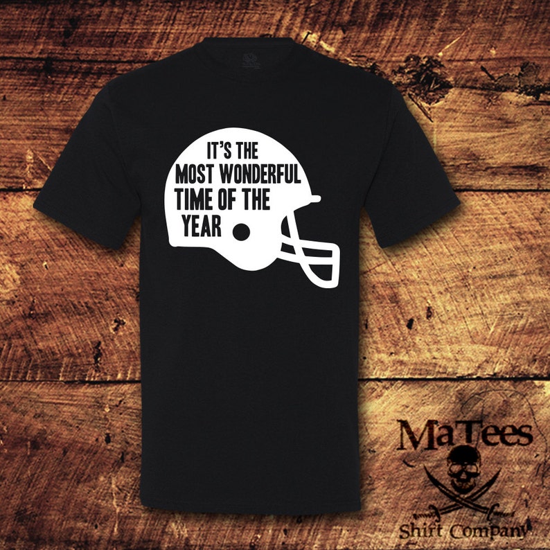 May include: Black t-shirt with white text that reads "It's the most wonderful time of the year" and a white football helmet graphic.