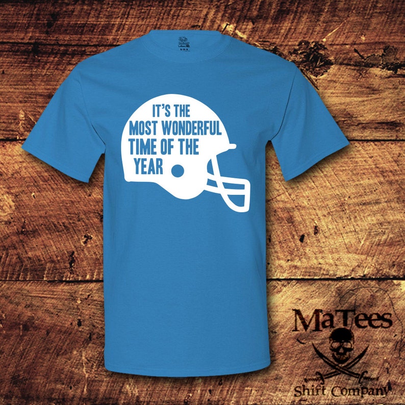May include: A light blue t-shirt with white text that reads "It's the most wonderful time of the year" and a white football helmet graphic.
