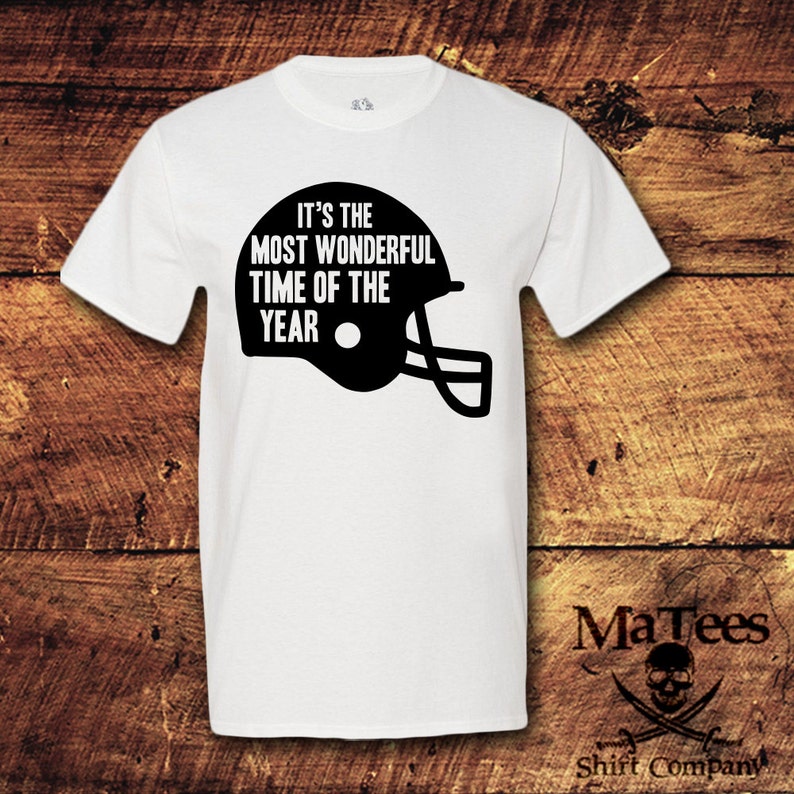 May include: White t-shirt with black text and a football helmet graphic. The text reads "It's the most wonderful time of the year."