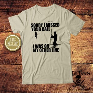 May include: A light tan t-shirt with the text "SORRY I MISSED YOUR CALL I WAS ON MY OTHER LINE" in black. The graphic shows a fisherman and a fish. The shirt also has the text "Shirt Company" and the "[a Tees" logo.