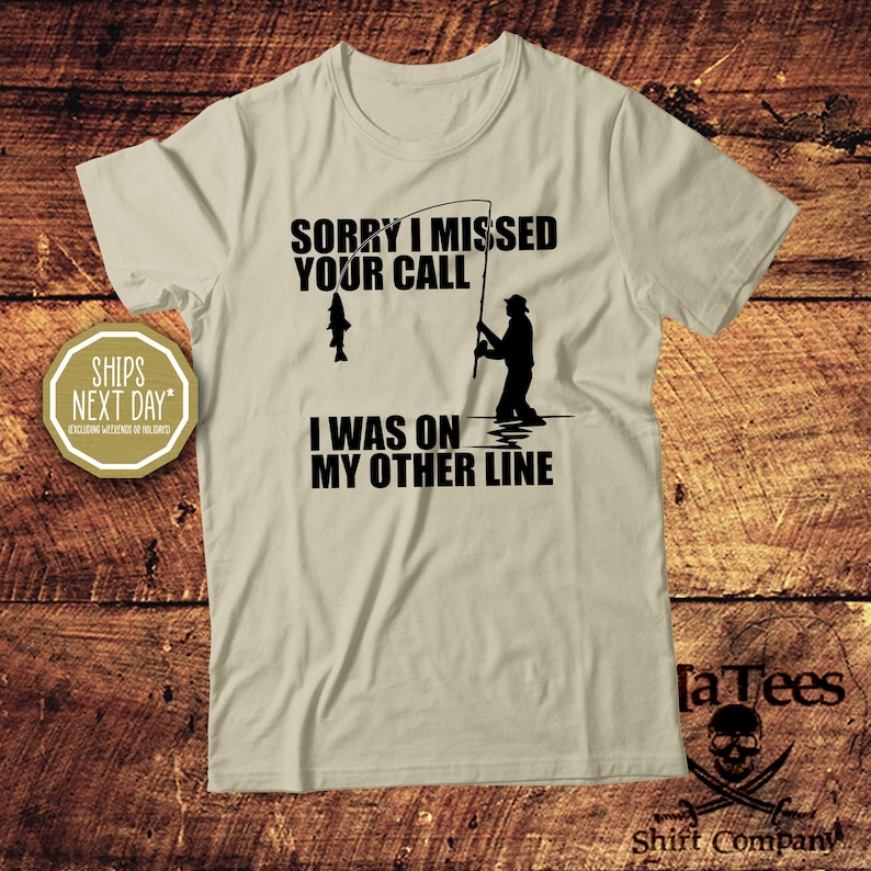 May include: A light tan t-shirt with the text "SORRY I MISSED YOUR CALL I WAS ON MY OTHER LINE" in black. The graphic shows a fisherman and a fish. The shirt also has the text "Shirt Company" and the "[a Tees" logo.