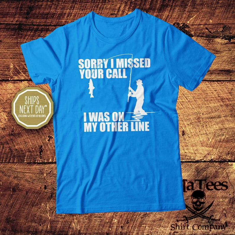 May include: A bright blue t-shirt with white text and graphics. The text reads "SORRY I MISSED YOUR CALL, I WAS ON MY OTHER LINE." A graphic shows a fisherman and a fish. The shirt is from "Ta Tees Shirt Company."