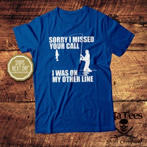 May include: A blue t-shirt with white text and graphics. The text reads "SORRY I MISSED YOUR CALL I WAS ON MY OTHER LINE." A graphic of a fisherman and a fish are also present. The shirt is from TaTees Shirt Company.