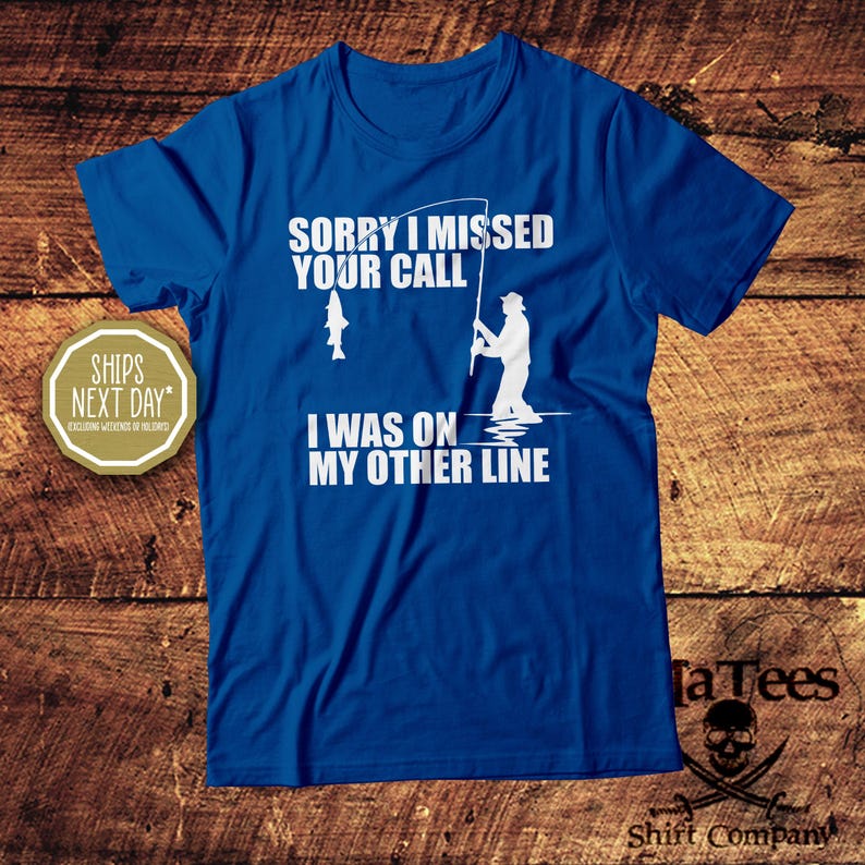 May include: A blue t-shirt with white text and graphics. The text reads "SORRY I MISSED YOUR CALL I WAS ON MY OTHER LINE." A graphic of a fisherman and a fish are also present. The shirt is from TaTees Shirt Company.