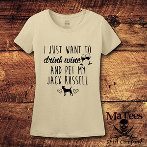May include: A beige t-shirt with the text "I just want to drink wine and pet my Jack Russell" in black. Two wine glasses clinking are above the text. A black silhouette of a Jack Russell terrier is below the text with two hearts on either side.
