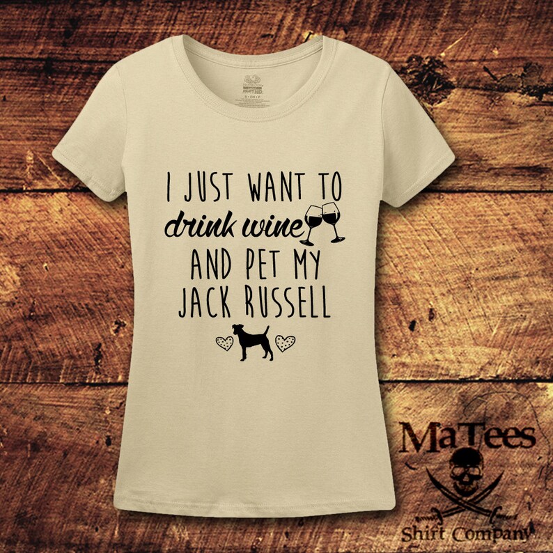 May include: A beige t-shirt with the text "I just want to drink wine and pet my Jack Russell" in black. Two wine glasses clinking are above the text. A black silhouette of a Jack Russell terrier is below the text with two hearts on either side.