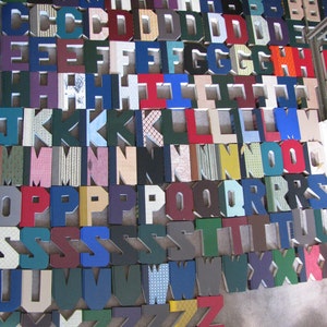 May include: A collection of colorful, three-dimensional letters arranged on a neutral surface. Each letter is a different color and pattern, including solid hues and patterned fabrics. The letters are made of cardboard or a similar material.
