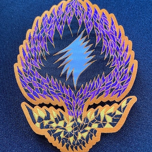 May include: A purple and orange embroidered patch with a stylized lightning bolt design. The patch is shaped like a flower with a pointed center and a rounded base.