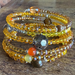 Puede incluir: Una pulsera de múltiples hilos con un diseño en espiral. La pulsera presenta una mezcla de cuentas ámbar, amarillas y marrones, con detalles metálicos plateados. Las cuentas son de varias formas y tamaños, creando un accesorio texturizado.