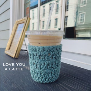 Könnte beinhalten: Ein türkisfarbener gehäkelter Kaffeetassenwärmer mit dem Text "Love You A Latte" auf einer dunklen Holzoberfläche.