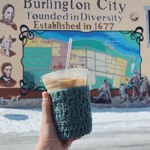 Peut inclure: Un manchon de tasse &agrave; caf&eacute; en crochet turquoise sur un gobelet de caf&eacute; glac&eacute;. L'arri&egrave;re-plan est une fresque de Burlington City, New Jersey, avec le texte "Burlington City Founded in Diversity Established in 1677".