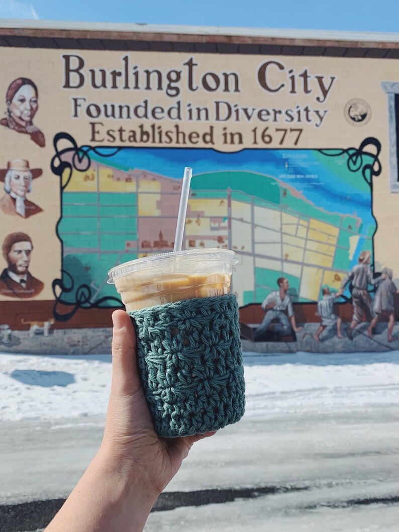 Peut inclure: Un manchon de tasse &agrave; caf&eacute; en crochet turquoise sur un gobelet de caf&eacute; glac&eacute;. L'arri&egrave;re-plan est une fresque de Burlington City, New Jersey, avec le texte "Burlington City Founded in Diversity Established in 1677".