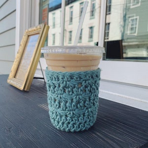 Peut inclure: Un manchon de tasse en crochet turquoise avec un gobelet en plastique transparent contenant une paille et du caf&eacute; glac&eacute;. La tasse est pos&eacute;e sur une surface en bois fonc&eacute;.