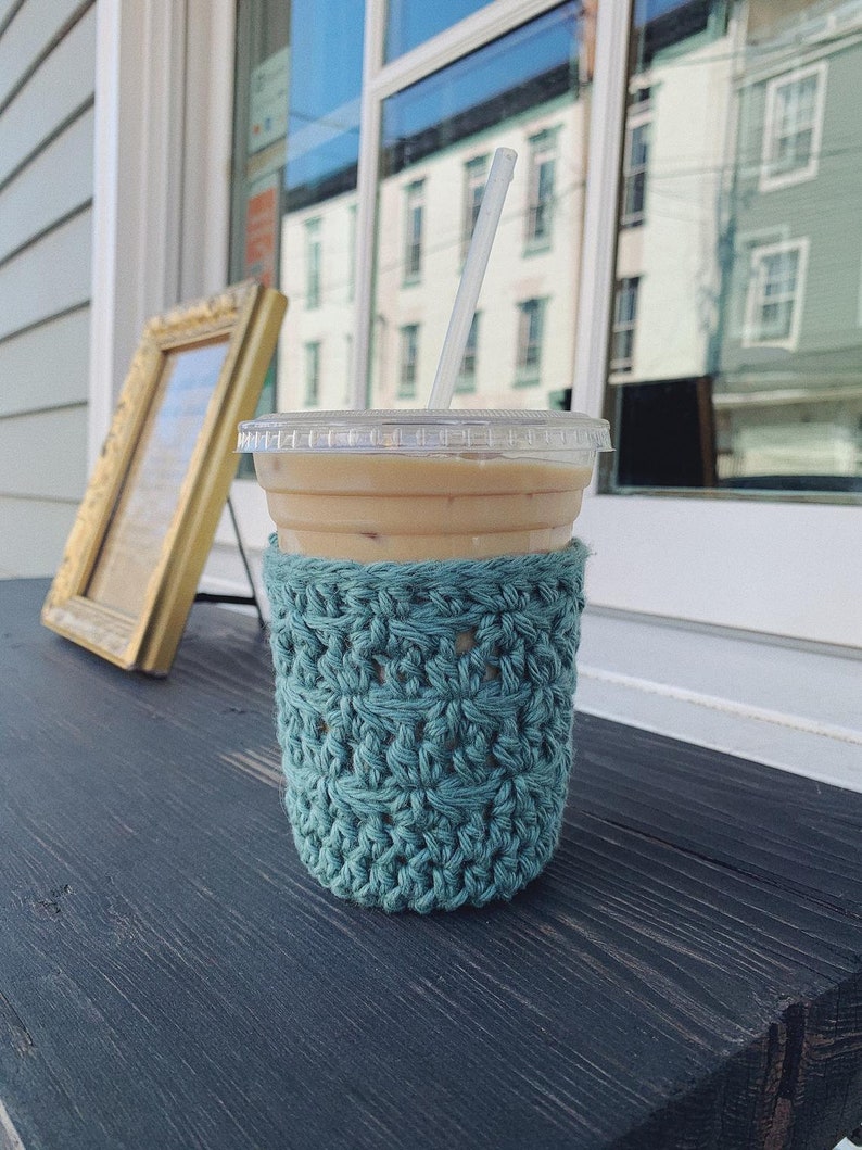 Peut inclure: Un manchon de tasse en crochet turquoise avec un gobelet en plastique transparent contenant une paille et du caf&eacute; glac&eacute;. La tasse est pos&eacute;e sur une surface en bois fonc&eacute;.