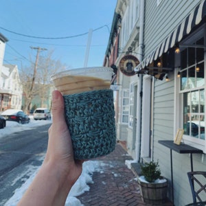 Peut inclure: Un gros plan sur un caf&eacute; glac&eacute; dans un gobelet en plastique transparent avec un manchon en crochet bleu. Le caf&eacute; est surmont&eacute; d'une paille.