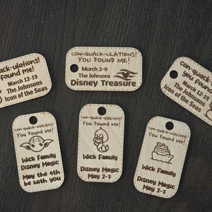 Puede incluir: Seis etiquetas de madera con un dise&ntilde;o caprichoso. Cada etiqueta presenta un personaje y un texto diferentes, incluyendo "con-quack-ulations! You Found Me!" y "Wick Family Disney Magic".